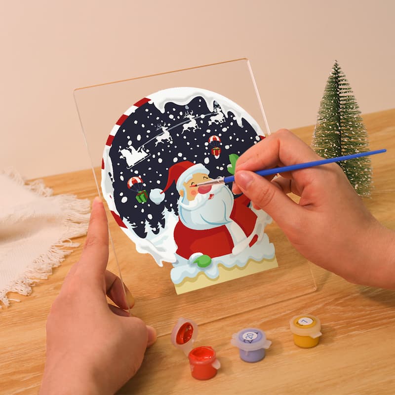 Person painting a Christmas-themed picture on a clear acrylic block with small paint pots on a wooden table.