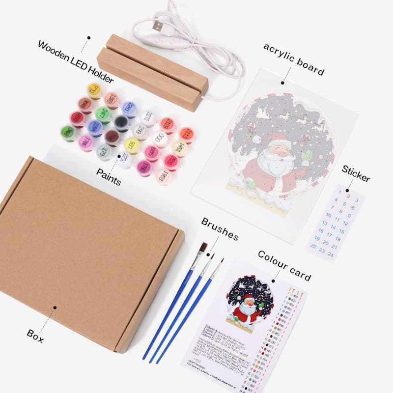 A flat-lay view of a Christmas Santa painting kit showing all its components: a brown cardboard box, wooden LED light holder, USB charging cable, acrylic board, paint pots, three blue-handled brushes, a sticker, and instruction cards, all labeled with text on a white background.