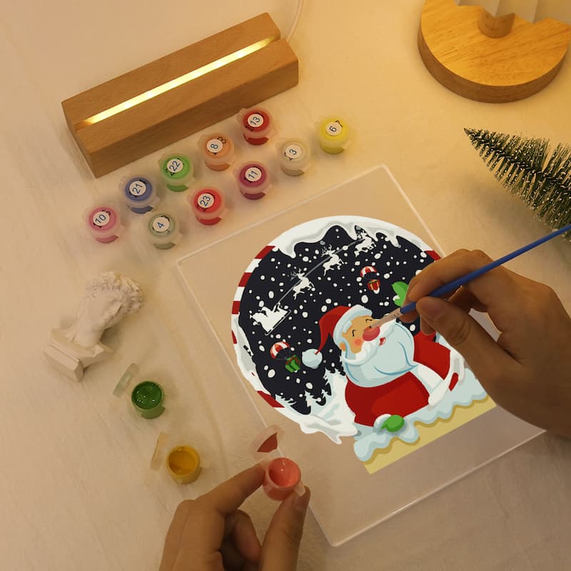 A person's hands are painting a Santa Claus scene on a clear acrylic panel using a blue paintbrush, surrounded by colorful paint pots, a wooden light box, and a small green tree on a light wood surface.
