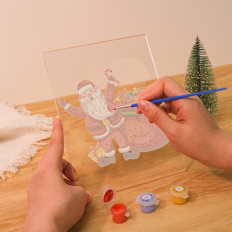 A person's hands are shown painting a Santa Claus design on a clear acrylic panel, using a small blue-handled paintbrush. Small paint pots are placed on a wooden table next to the panel, with a miniature Christmas tree in the background.