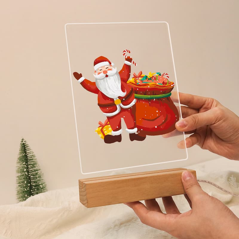 Hand holding a transparent acrylic plaque with a Santa Claus design on a beige background.