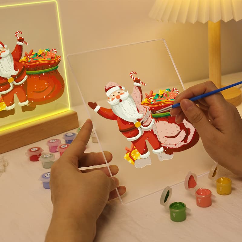 A person is painting a Christmas-themed panel with a Santa Claus and a sack of candy using a blue paintbrush. The scene includes small paint pots on a table, a lit lamp in the background, and a finished glowing panel reflected on the left.
