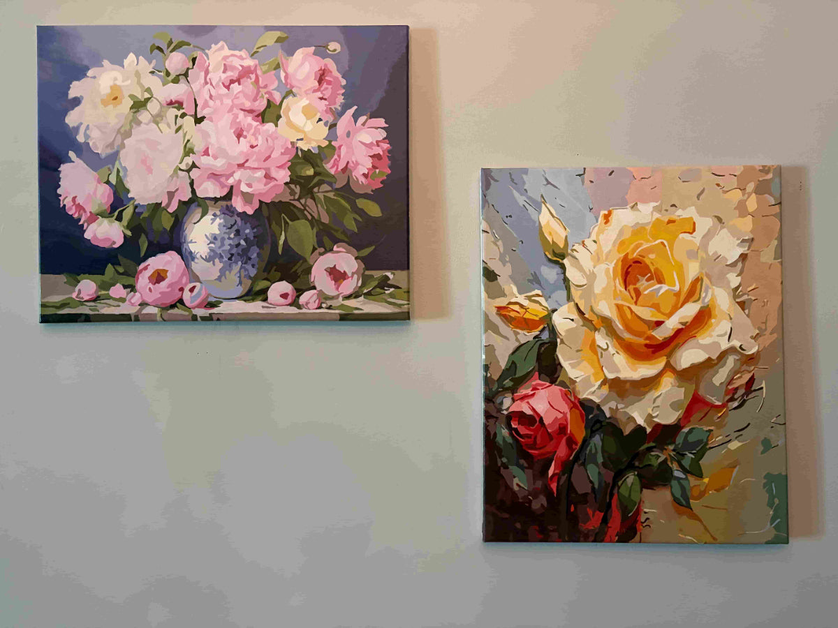 Two square floral canvas paintings hanging on a light gray wall. The left painting shows pink and white flowers in a blue-and-white vase against a dark blue background.