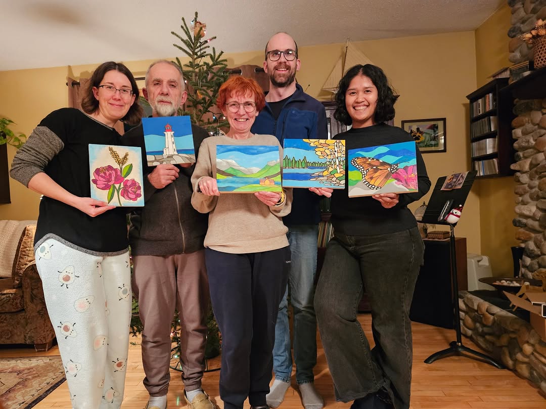 Happy Canadian family proudly displaying their finished Paint By Numbers artworks in a cozy living room. Each person holds a vibrant painting, including flowers, landscapes, a lighthouse, and a butterfly, capturing a joyful and creative moment together.