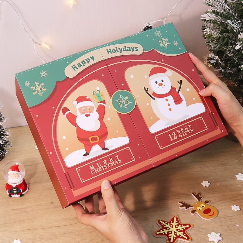 Hands holding a red and green Christmas-themed gift box featuring Santa Claus and a snowman illustration with text “Merry Christmas” and “12 Best Gifts” on the front, placed on a wooden table with festive decorations.