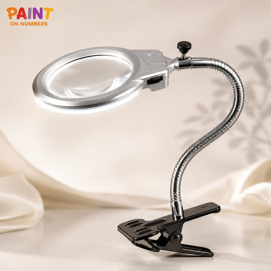 A silver and black clip-on magnifying lamp with a flexible gooseneck arm and illuminated ring, shown on a soft beige draped fabric background. The 'PAINT ON NUMBERS' logo is visible in the top-left corner.