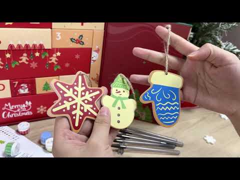 Hands holding painted wooden Christmas ornaments shaped like a star, snowman, and mitten in front of a festive red gift box with numbered drawers from the 12-Day Christmas Crafter’s Gift Box by Paint On Numbers.